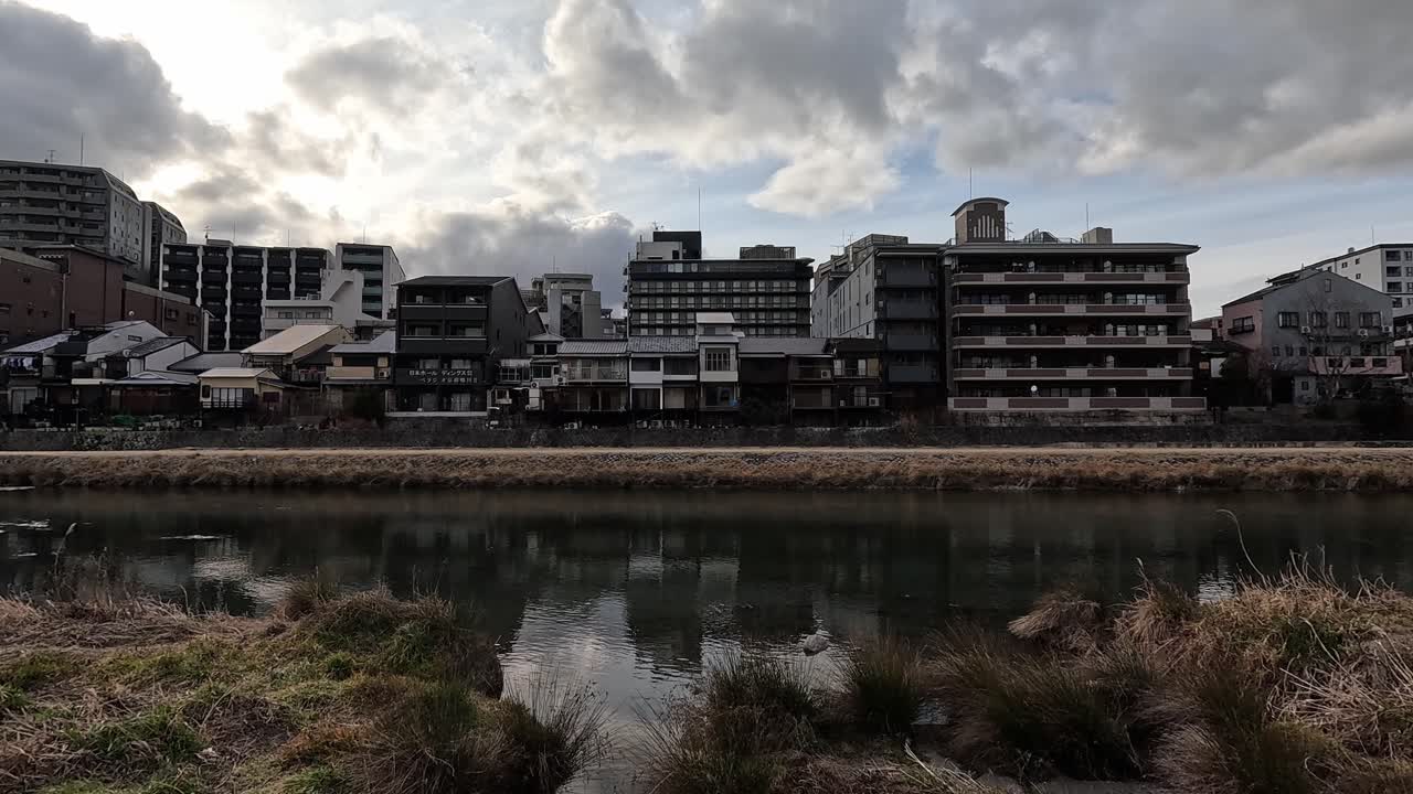 Urban Waterfront View of Kyoto’s Kamo River with Reflections and Traditional Japanese Architecture under Cloudy Sky