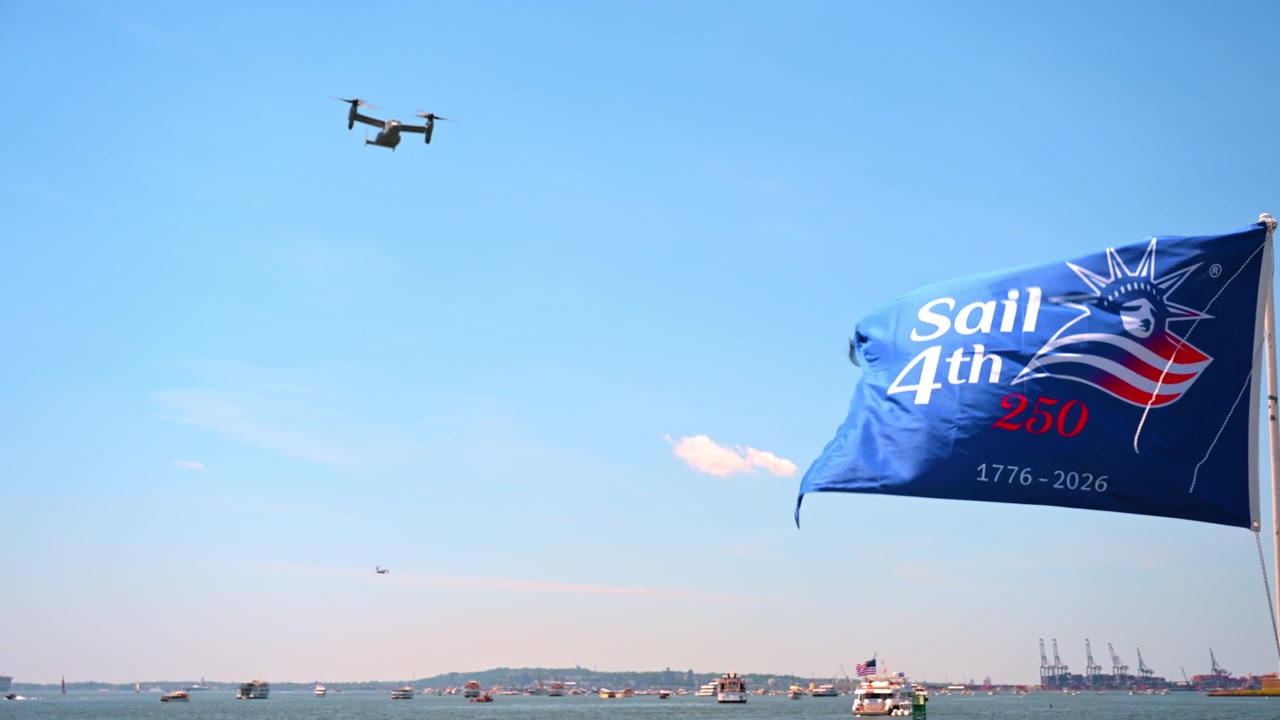 Sail4th 250 flag and tiltrotor over New York Harbor. Boats fill the sunlit water beneath a clear blue sky on Independence Day.