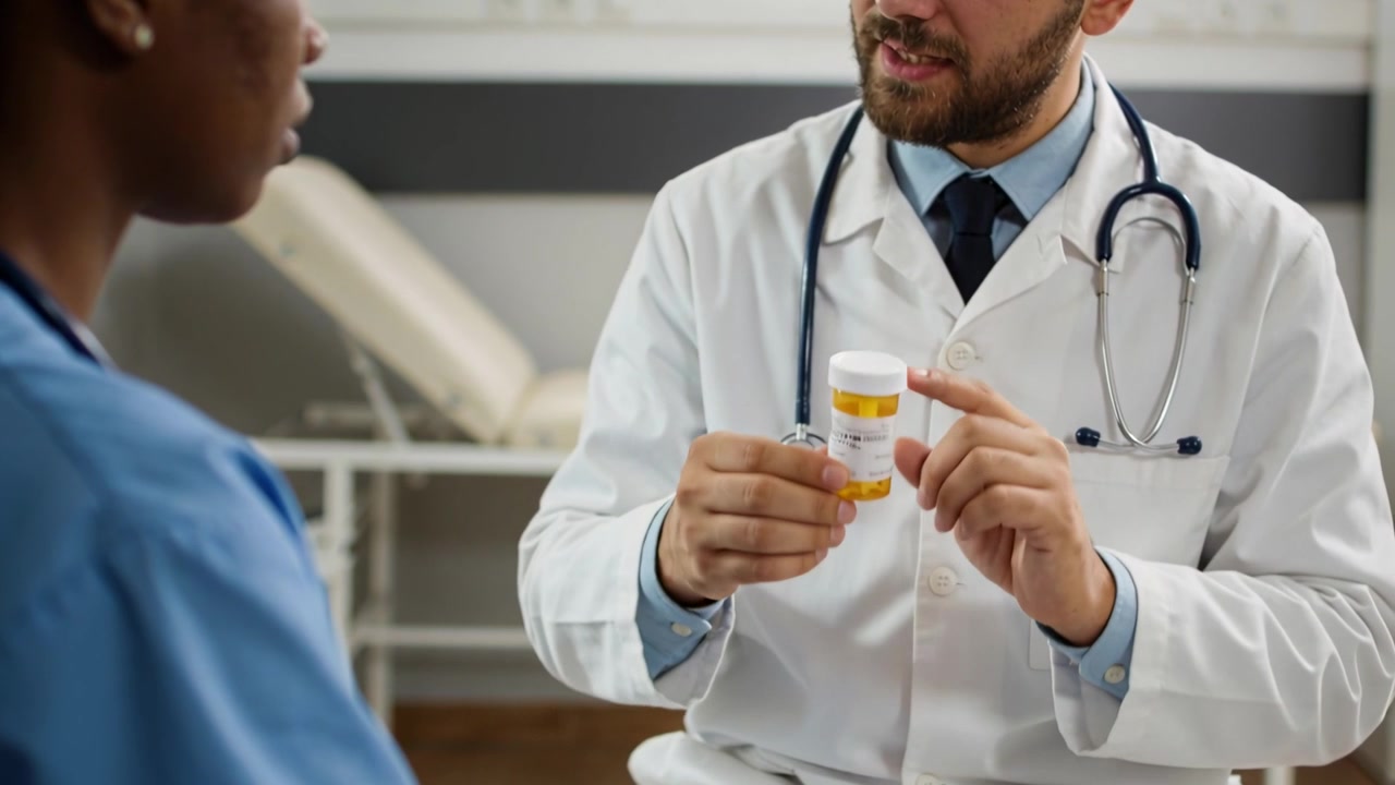 Segurando frasco de comprimidos, médico de jaleco e estetoscópio mostrando rótulo explicando a dose na clínica, com ouvinte de uniforme médico.