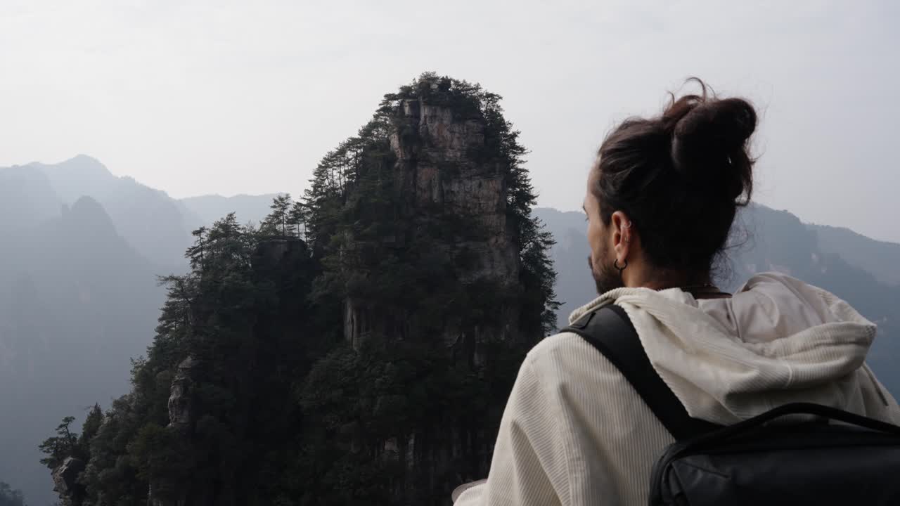 A man with a backpack gazes at a towering sandstone peak in Zhangjiajie, surrounded by misty mountains and dense forest.