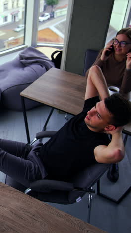 people working in the office having a break from work. man sitting in chair with legs on the desk and looking at window. woman speaks on phone. top view. vertical video