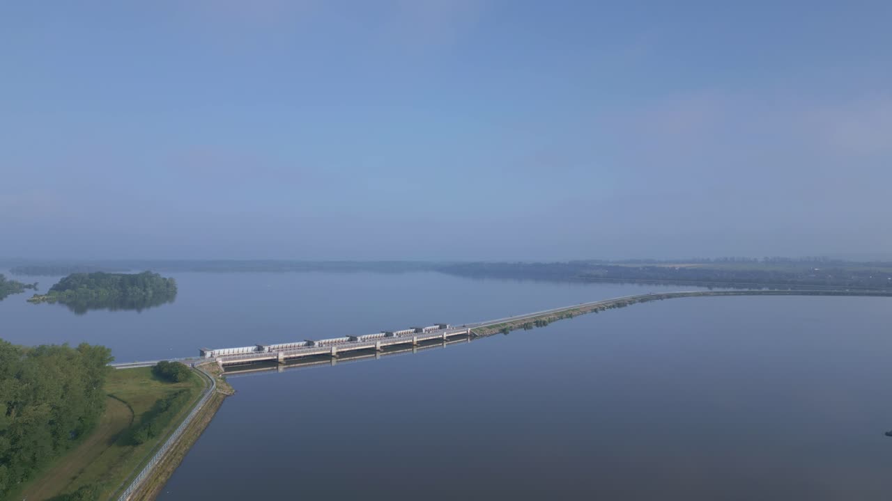 Aerial view of the long dam over the calm water surface of the Věstonice reservoir with surrounding greenery and clear sky