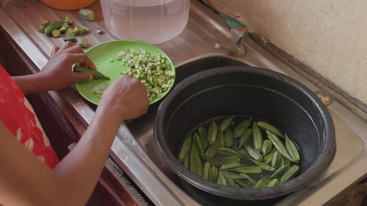 Black woman shelling peas at sink, transferring fresh peas into green bowl while empty pods collect in dark pot, warm natural light, closeup on careful hands, slow domestic routine, textured counter
