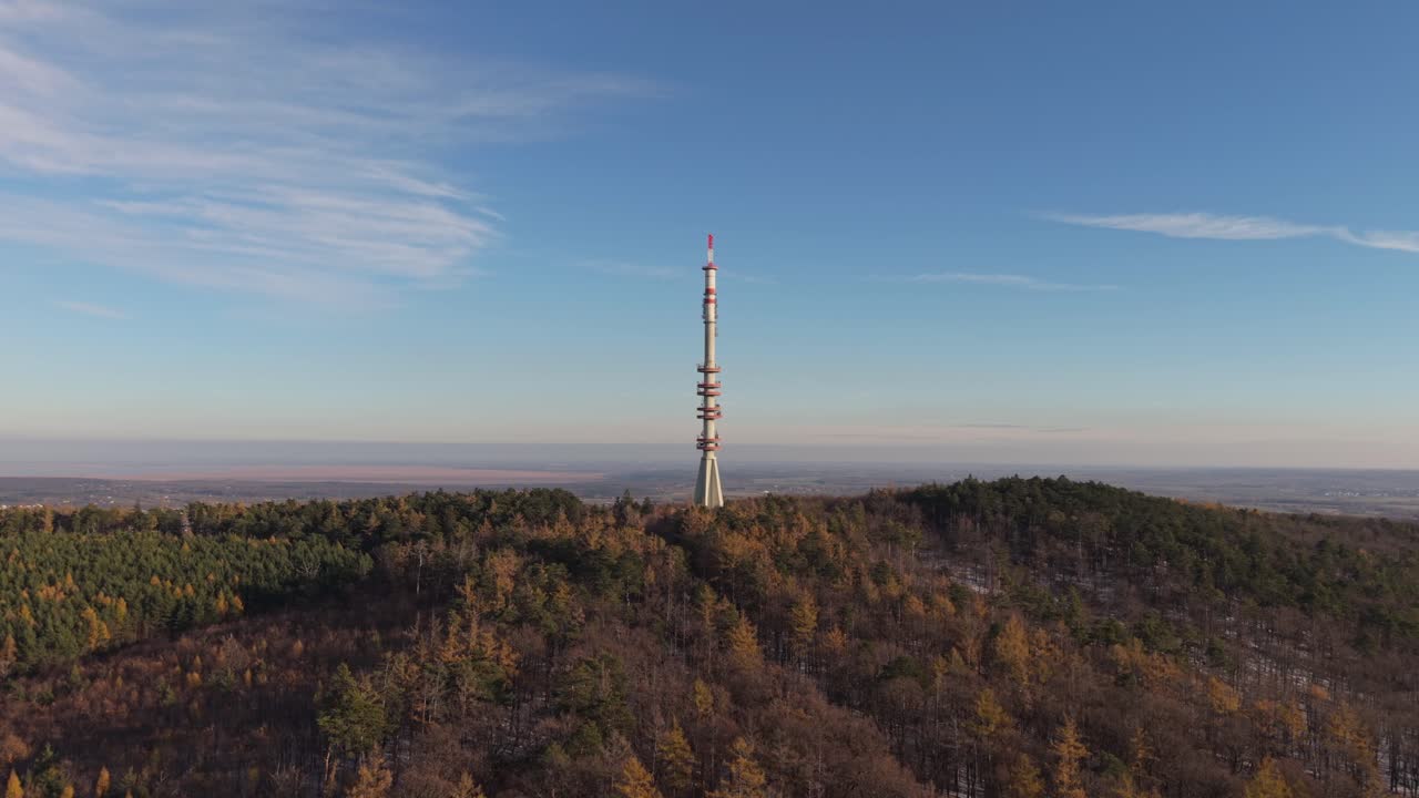 An aerial video of a tall telecommunication tower rising above a forested hillside. The scene captures the contrast between modern infrastructure and the natural landscape under a clear blue sky