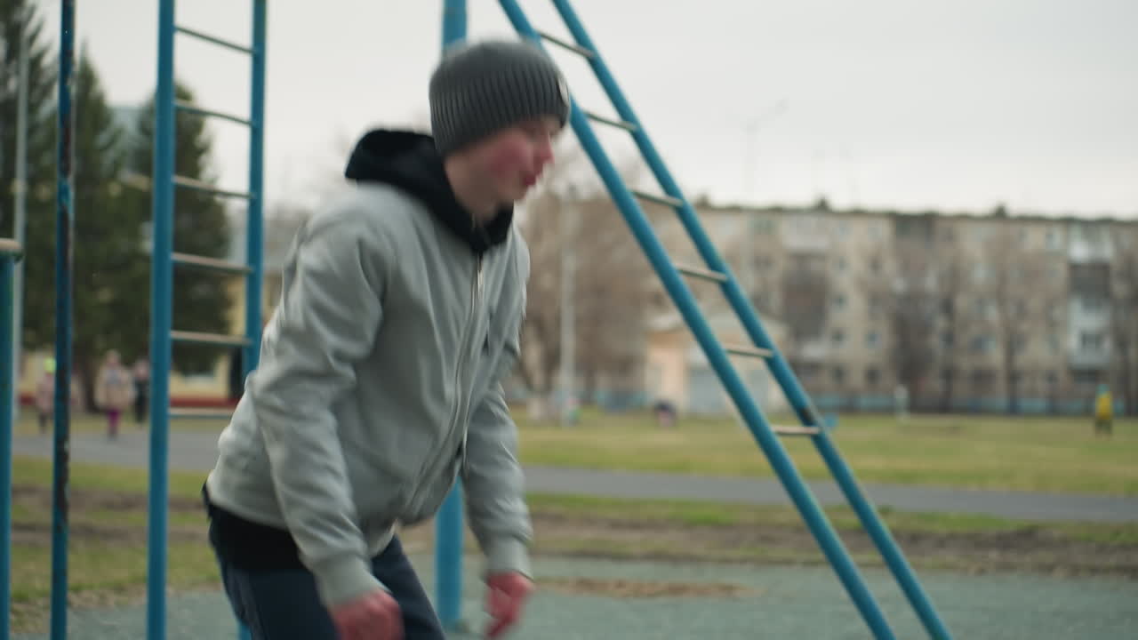A young boy performs a workout with hands stretched out close to an iron pole, in a stadium with a yellow building in the background, a blurred view of people walking near the building