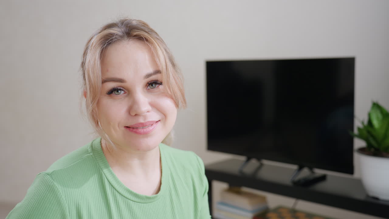 Cute woman touches kid gently while looking with smile toward camera showing warmth love and care in bright room with television books and green plant in background
