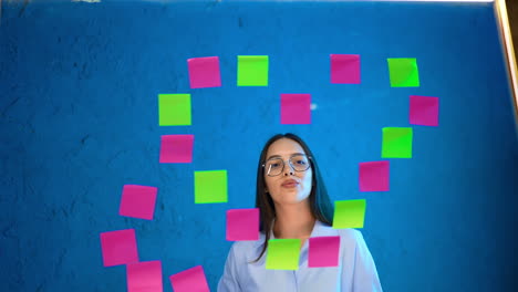 smiling caucasian lady in glasses puts colorful sticky notes on the glass wall. placing memos on the board. business concept, list to do.