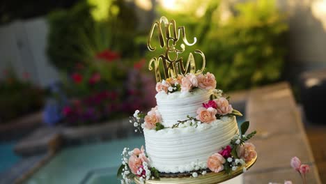 Shot-of-a-beautifully-decorated-floral-wedding-cake-sitting-on-a-pedestal-right-next-to-a-pool-during-a-summer-wedding-reception