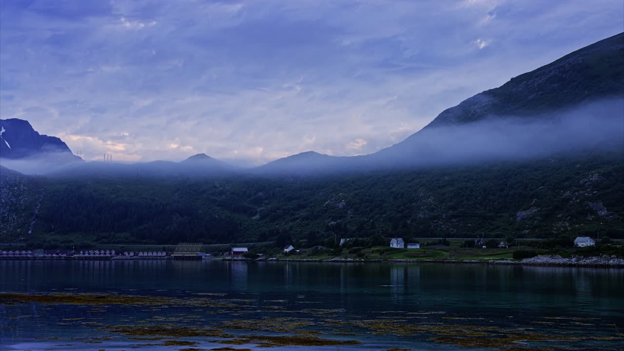 Foggy, lush, green mountains with busy, winding road, houses and cabins by the water during low tide on a bright Arctic summer night. Wide angle panning time-lapse. Lofoten, Northern Norway