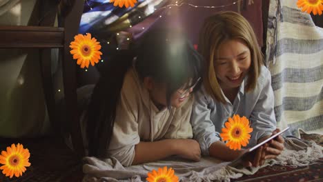 animation of orange flowers over happy asian mother and daughter using tablet at home