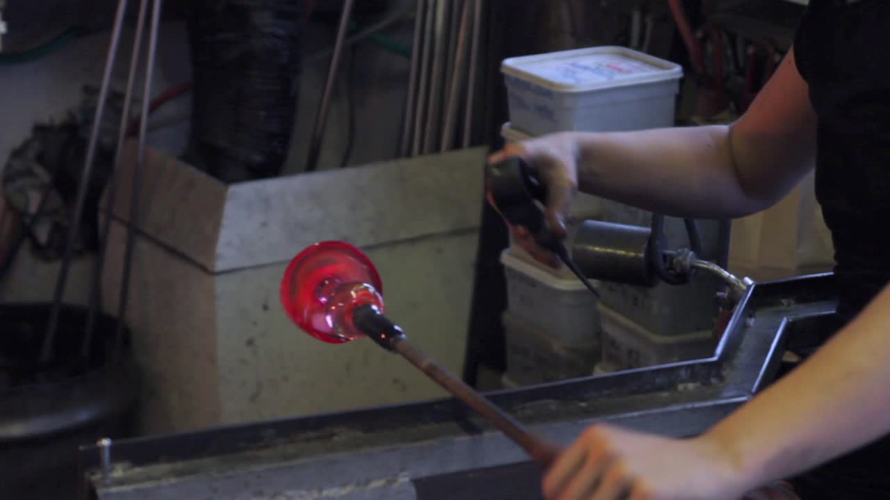 Mid shot of female glass worker, turning and shaping hot glass with crimps in a dark forge