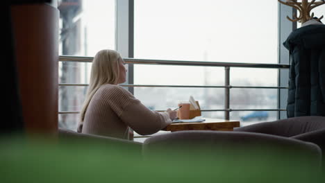 side view of elegant student thoughtfully writing, pauses to rest hand on jaw, continues writing with coffee cup on table and sweater on cloth rack in busy urban winter setting