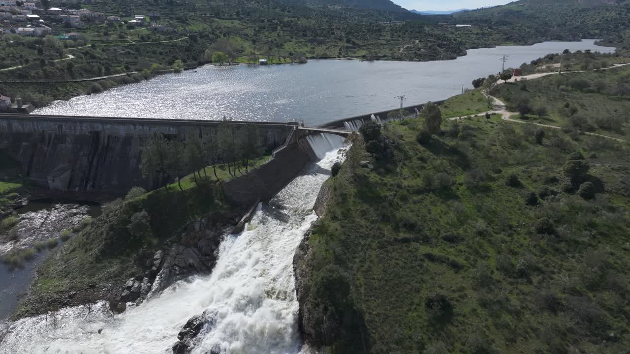 Slow motion drone shot of a dam at full capacity, with its side overflow releasing a massive amount of water, forming a white cascade as it crashes and foams in a powerful descent.