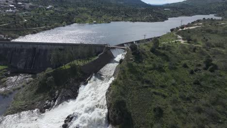slow motion drone shot of a dam at full capacity, with its side overflow releasing a massive amount of water, forming a white cascade as it crashes and foams in a powerful descent.