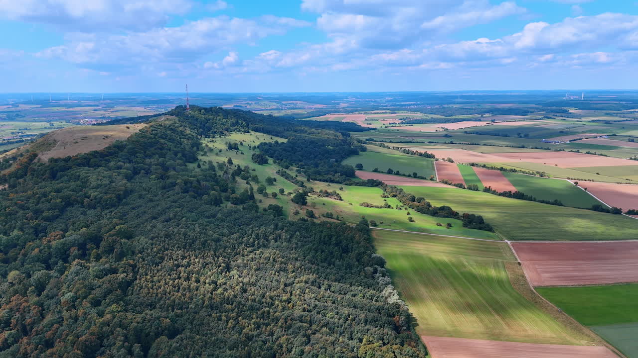 Een uitzicht op uitgestrekte velden en groene heuvels. Het landschap omvat stukken bruin en groen land. In de verte staat een kleine heuvel met een toren erop.