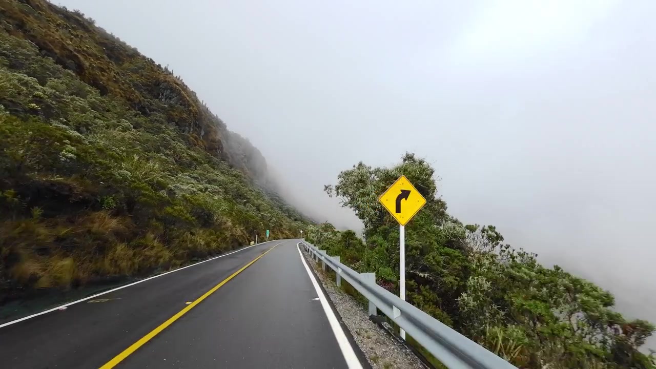 Una POV del veicolo segue la strada Murillo–Manizales accanto a un ripido pendio coperto di frailejones, mentre una densa nebbia bianca di montagna limita la vista in lontananza