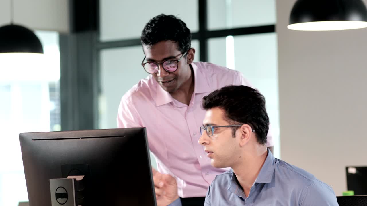 two young male office employees, workers or office staff members are discussing and interacting in front of computer monitor
