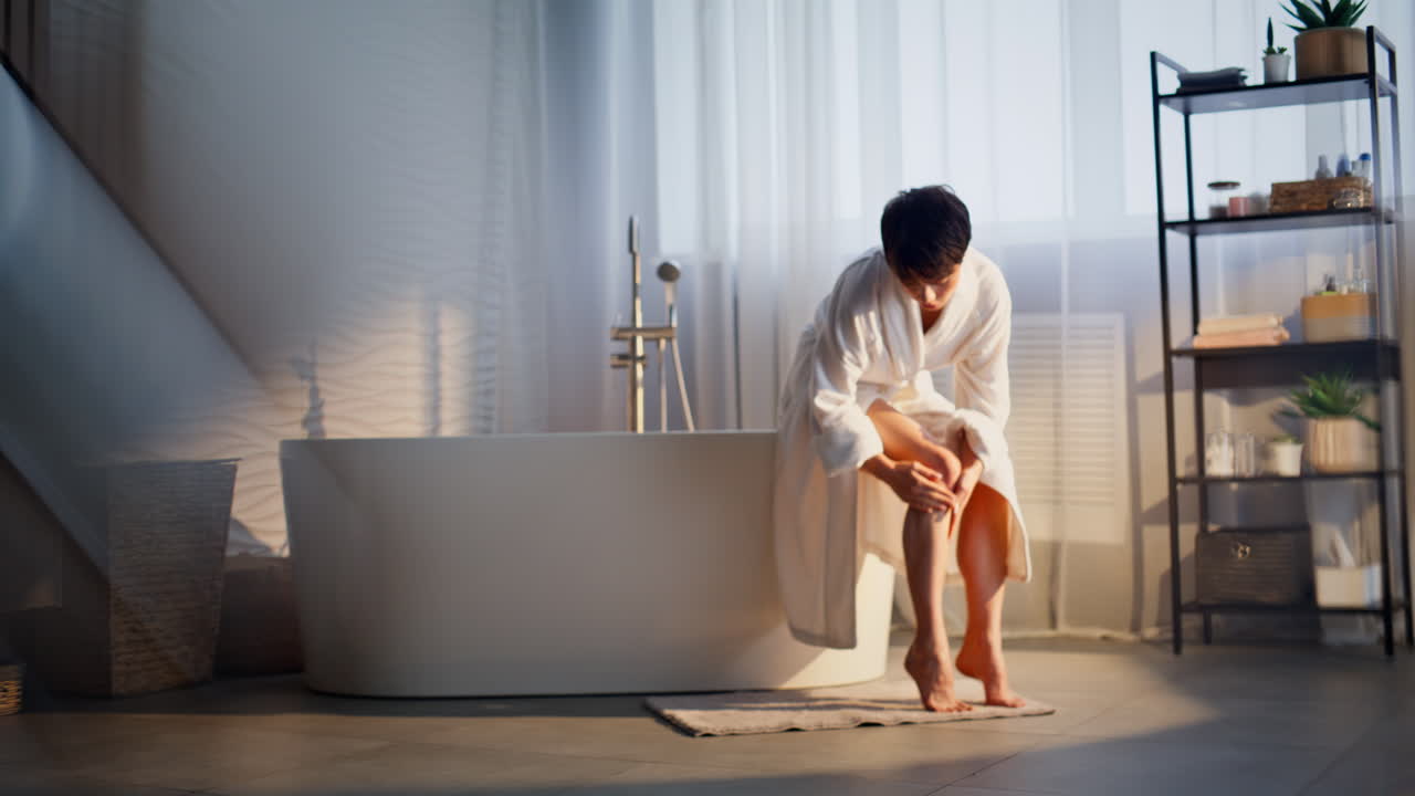 Woman putting legs cream indoors. Serene white gown model resting weekend alone