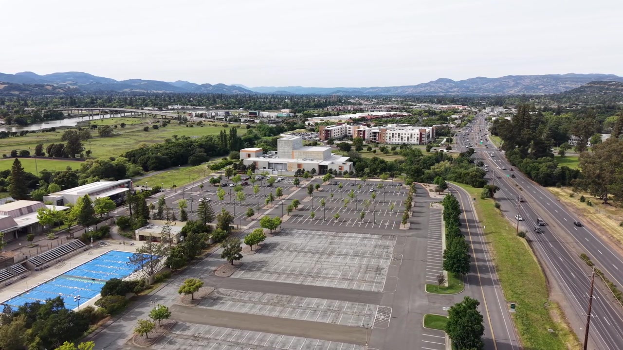 Vista aérea panorâmica do campus da Napa Valley College com estacionamento vazio, piscina ao ar livre e rodovia adjacente com tráfego em movimento contra um cenário de encosta verde