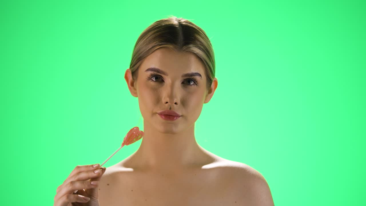 Static shot of a young pretty woman or model licking a heart shaped lollipop and giving an attractive look to the camera while enjoying the candy in front of green background