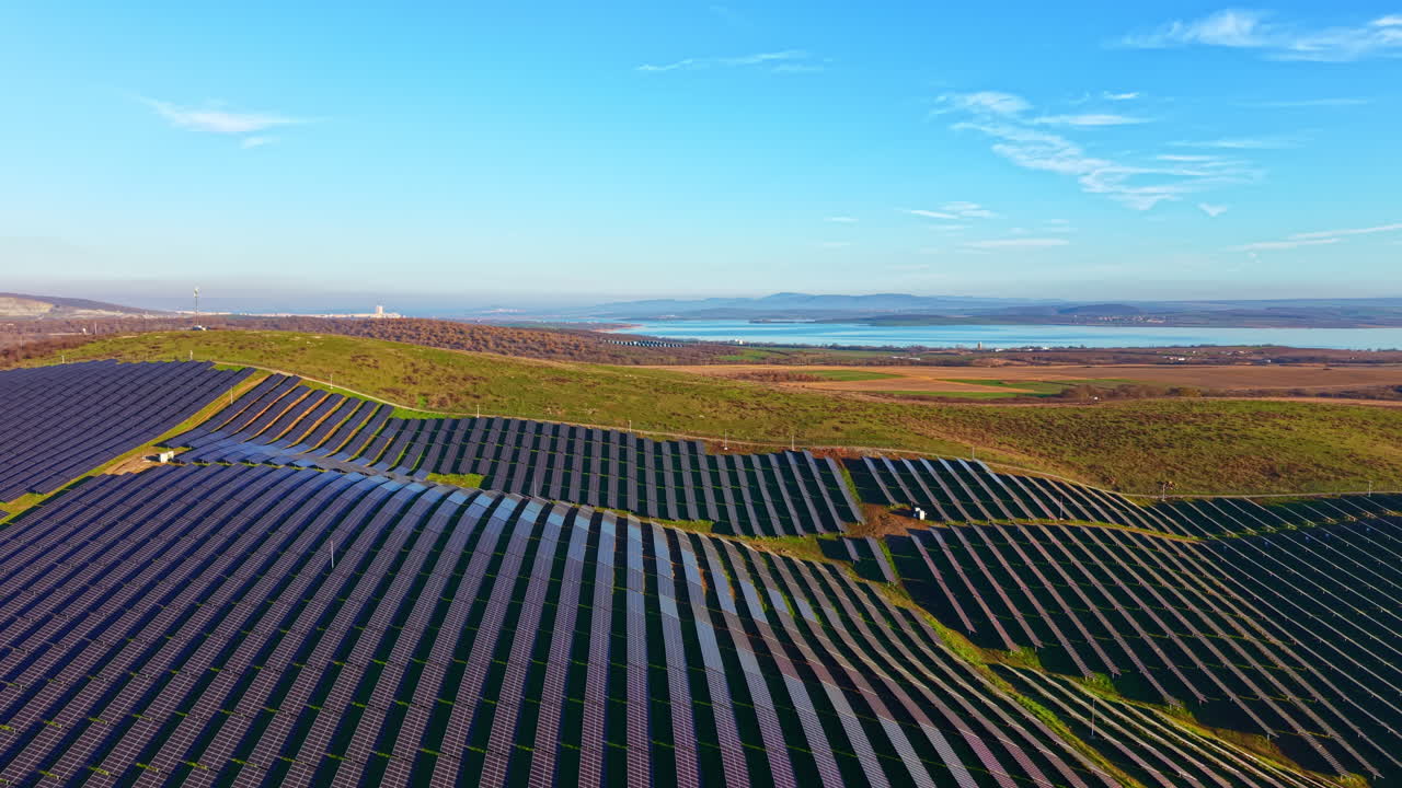 Store solcelleparker strækker sig over bakker under en lys blå himmel nær en fredfyldt sø i den gyldne time, hvilket viser vedvarende energi i harmoni med naturen.