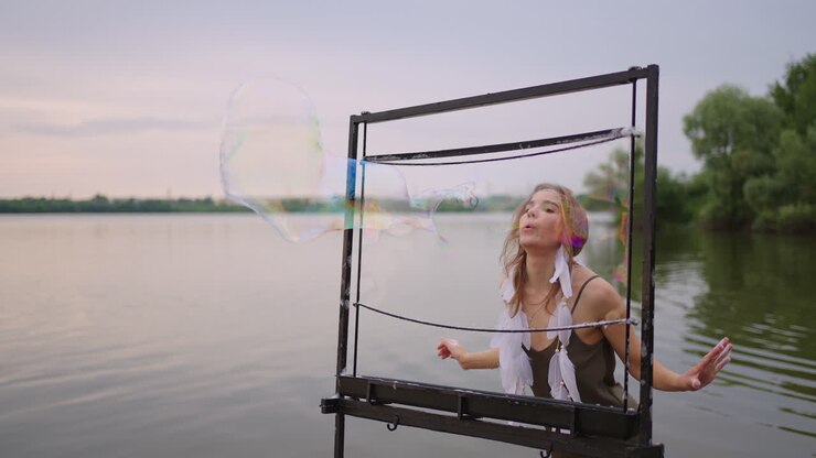 Una giovane artista donna soffia molte bolle di sapone, presentando uno spettacolo teatrale con una cornice al rallentatore al tramonto su un lago.