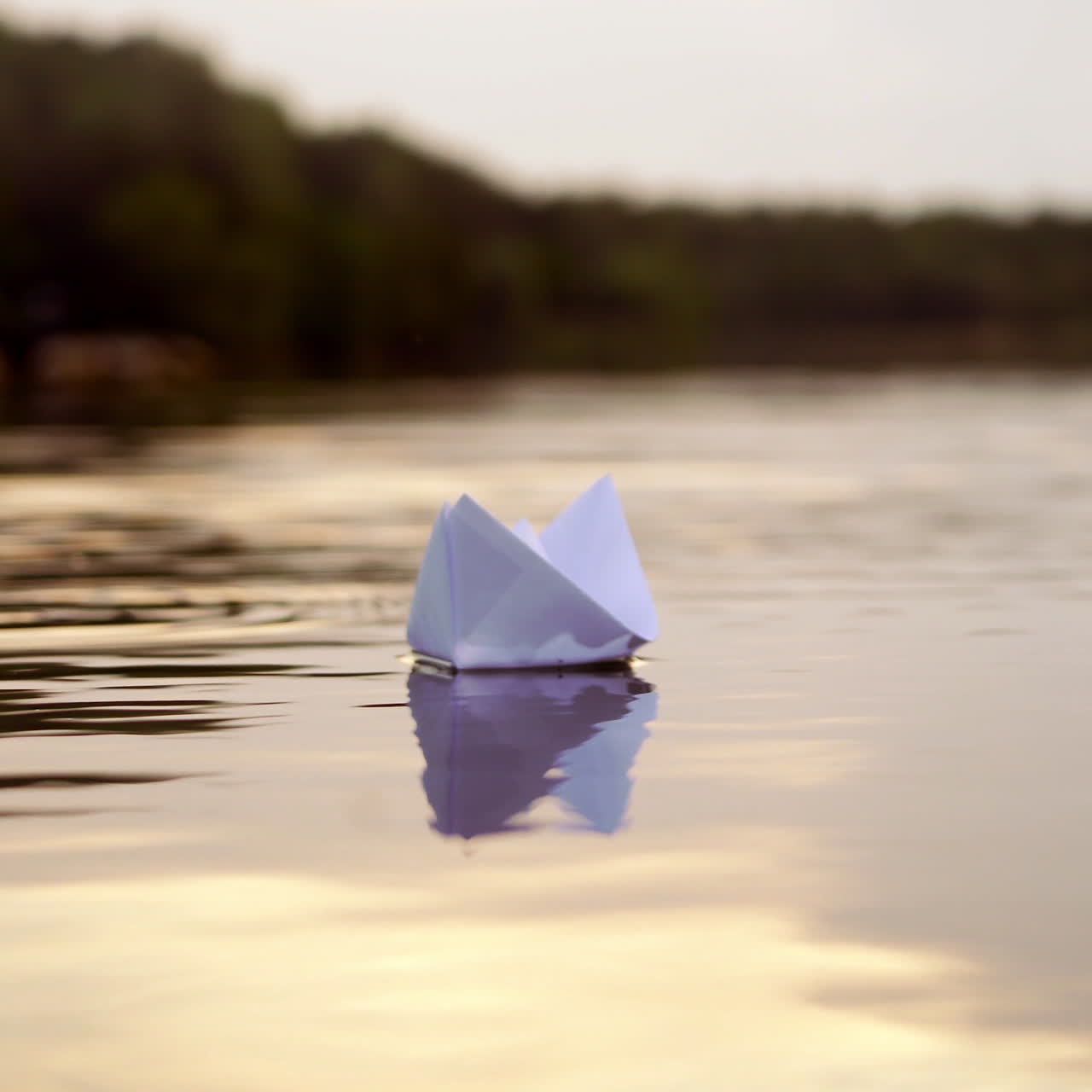 White paper boat is floating on the water near the forest background in the evening. Origami boat floats away into the distance along the river
