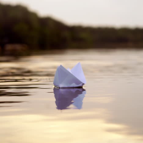 white paper boat is floating on the water near the forest background in the evening. origami boat floats away into the distance along the river