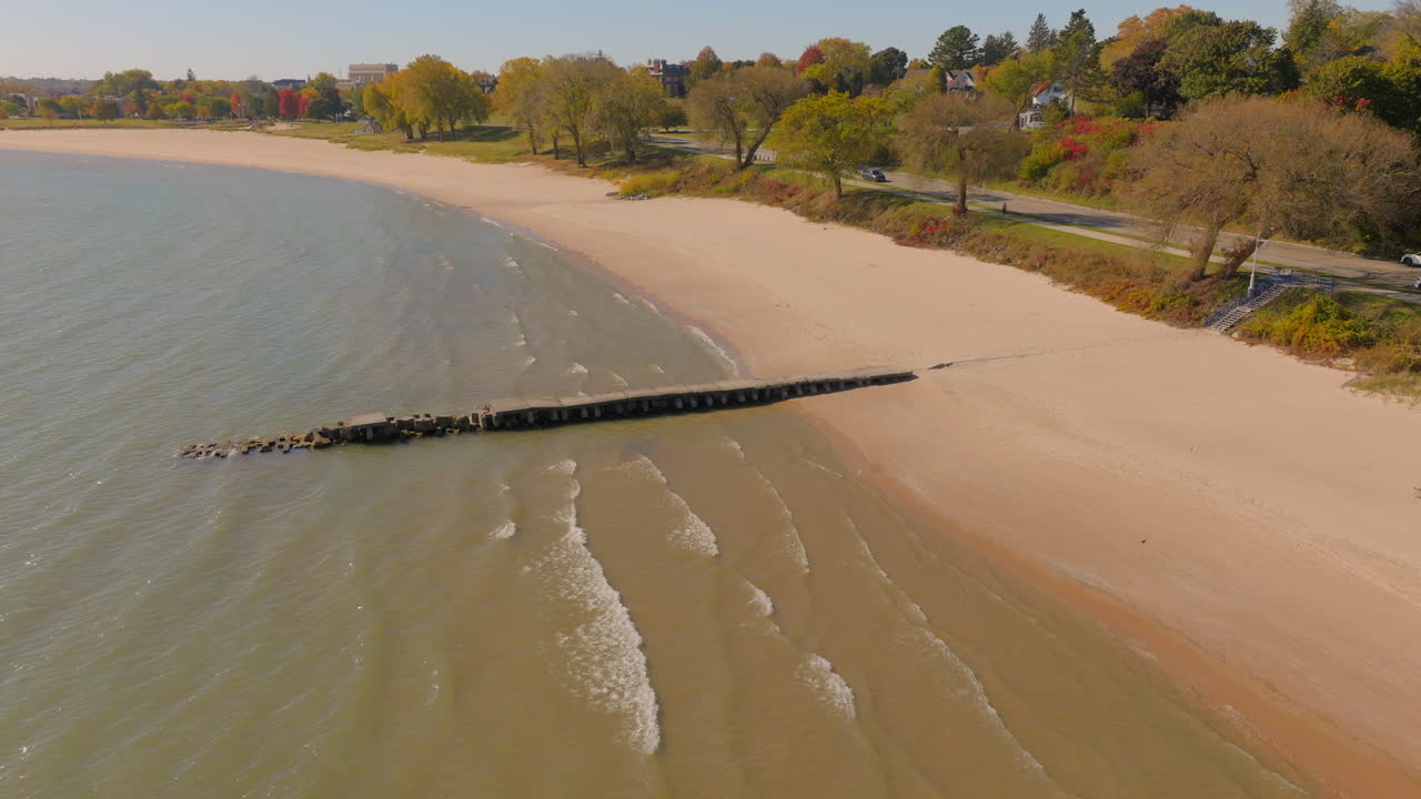 Drone aerial rising from Lake Michigan shoreline with concrete pier, revealing a charming Sheboygan, Wisconsin neighborhood of colorful trees and homes on a bright blue-sky day