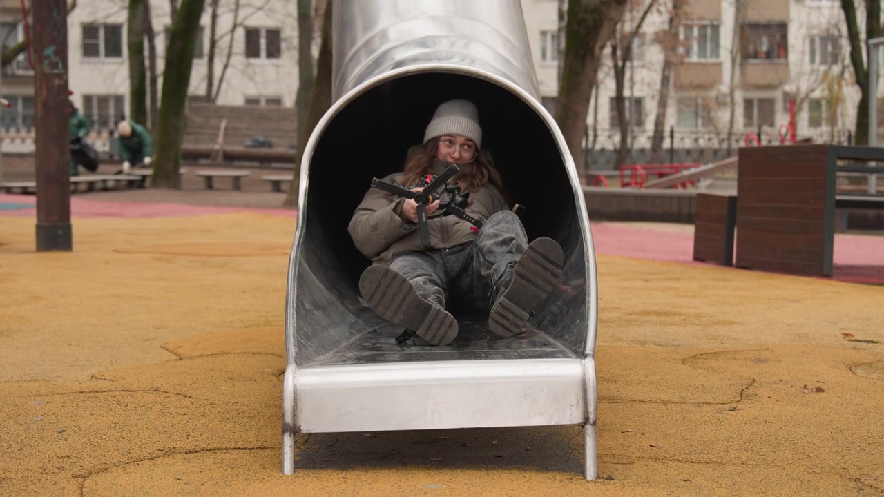 Adult woman inside metal slide playground curled in winter coat and beanie, clutching small bag, apartment buildings in background, rubber safety surface, muted palette, candid documentary style