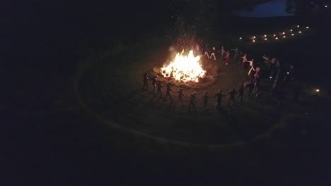 Crowd-of-people-dancing-around-bonfire-at-night,-aerial-orbit-view