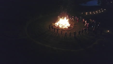 crowd of people dancing around bonfire at night, aerial orbit view