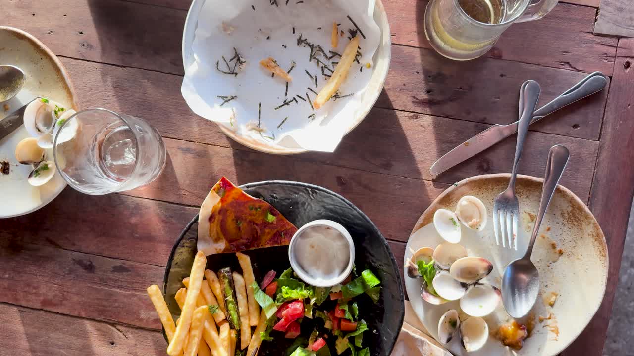 Aerial view of a vibrant meal with pizza, fries, clams, and salad on a wooden table under warm natural light