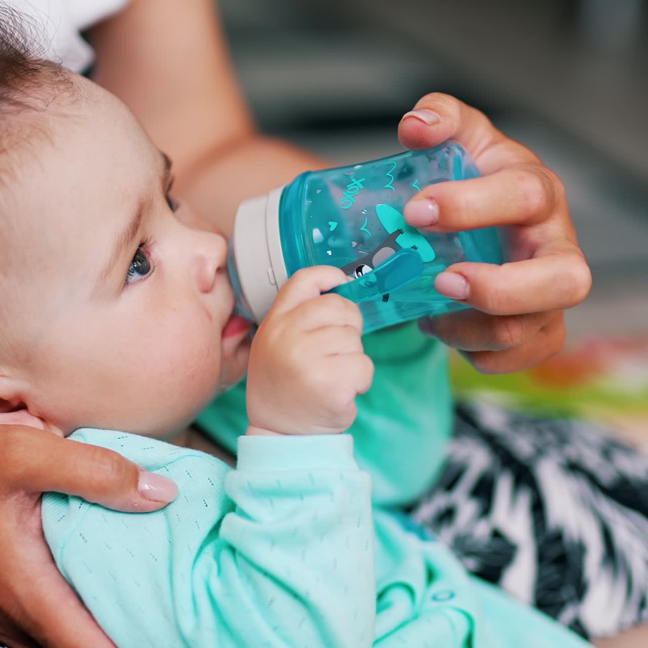 Cute boy half-sitting in mom's hands drinks water. Adorable baby with funny hair suckling bottle
