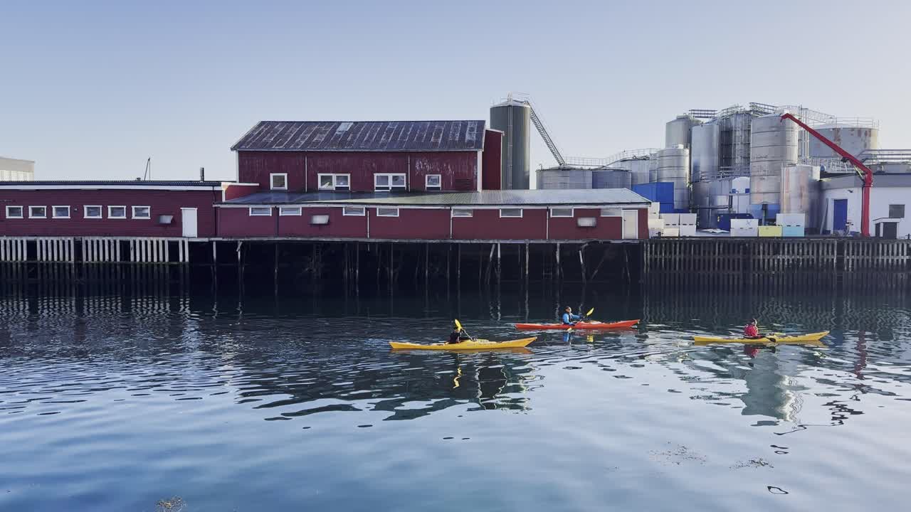 Kayakers paddle in calm waters near red buildings in Lofoten, clear sky, midday light, serene vibe, wide shot