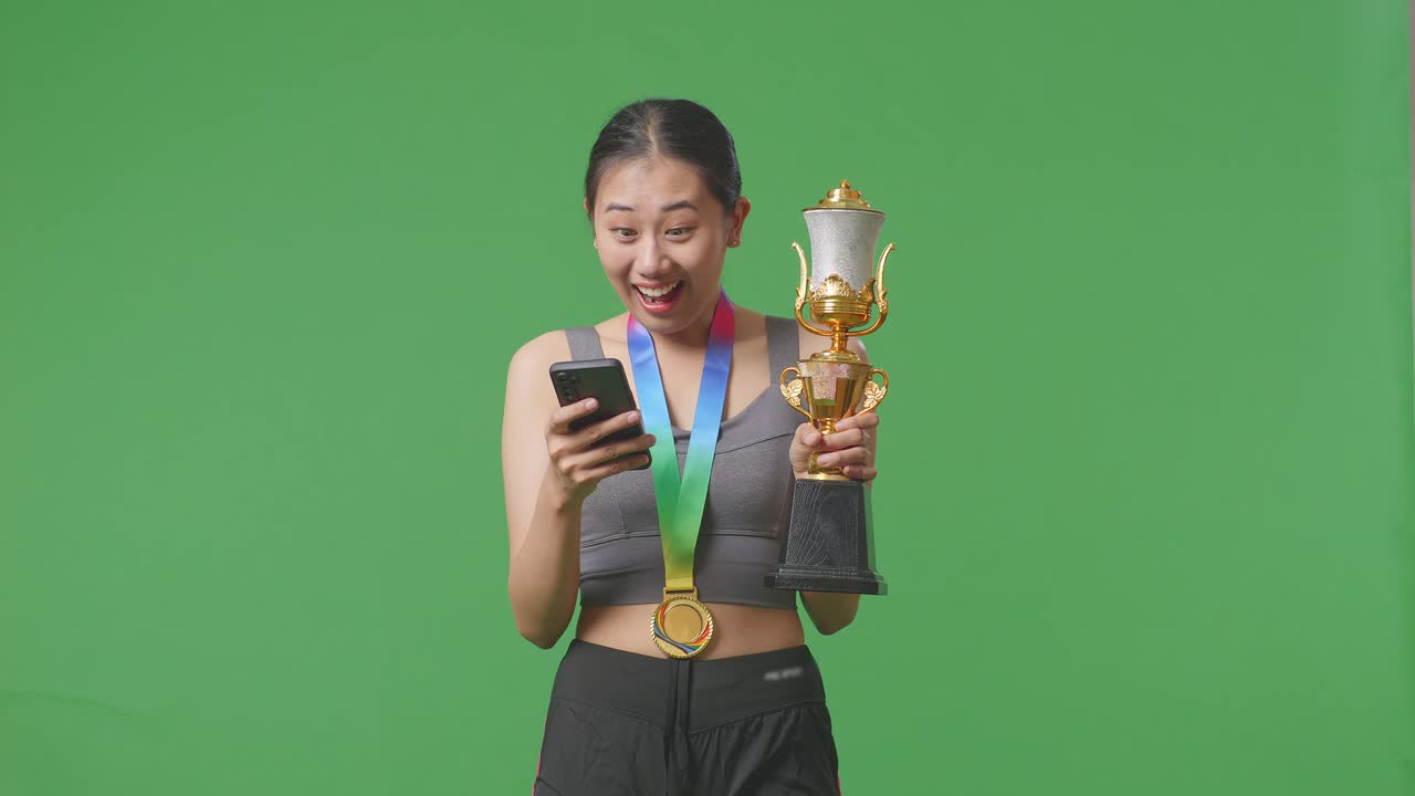 금메달과 트로피를 가진 아시아 여성이 스마트폰을 보고 스튜디오의 녹색 화면 배경에서 와우라고 말합니다.