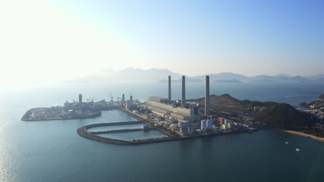 Drone-shot,-traveling-to-the-left,-of-a-big-coastal-coal-fired-power-plant-on-an-island-next-to-a-village-with-a-beach-during-a-beautiful-sunny-day-2