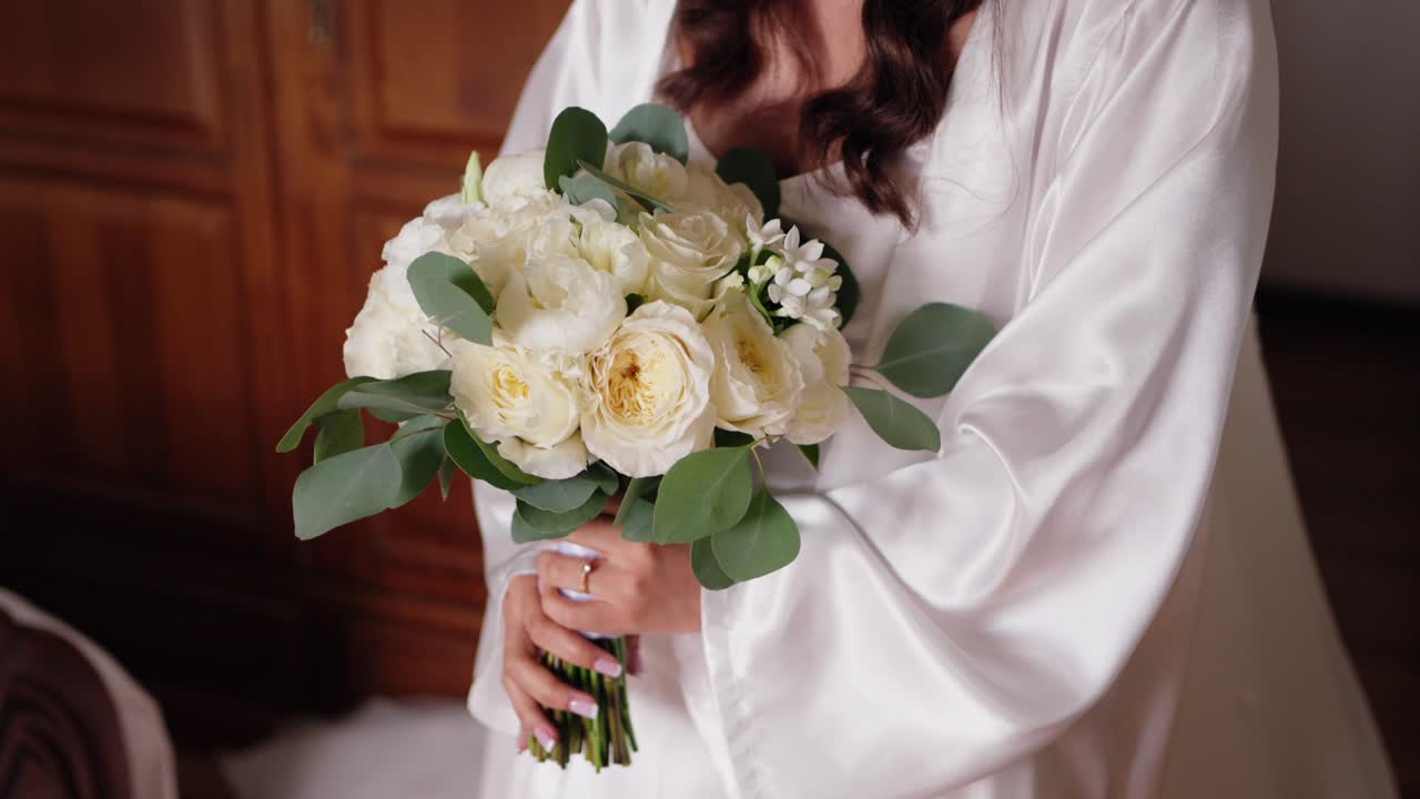 Sposa in vestaglia di seta che tiene un bouquet di rose bianche e foglie di eucalipto verdi.
