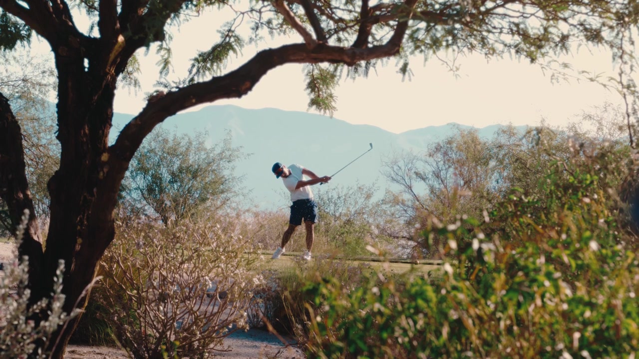 Scratch golfer swings framed through desert mesquite trees in ultra slow motion, hazy mountains beyond the branches, par 3 perfect swing on sunny summer day