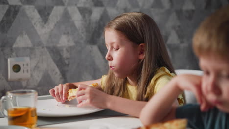 close up blur view of little boy sitting close to sister in yellow shirt eating cheesy pizza at dining table with glass of juice placed on table in bright room with patterned wall background indoors