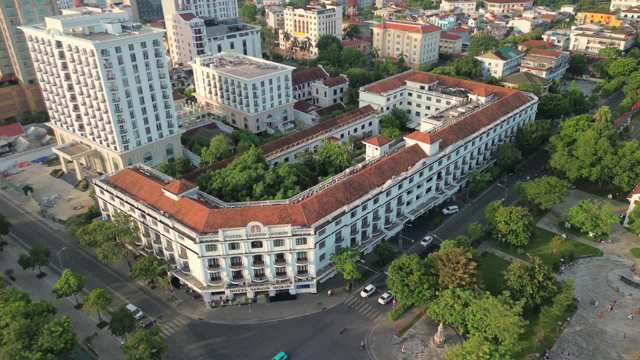 Foto de drone em ângulo alto do icônico edifício branco em estilo colonial com telhados de terracota no centro da cidade de Hue, cercado por árvores verdejantes e tráfego urbano