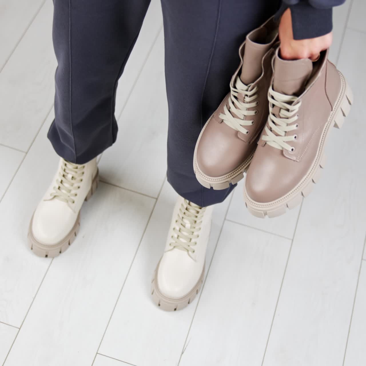 Female model wearing loose pants and white tractor sole boots holding another pair of beige boots in hands. View from above. White floor backdrop