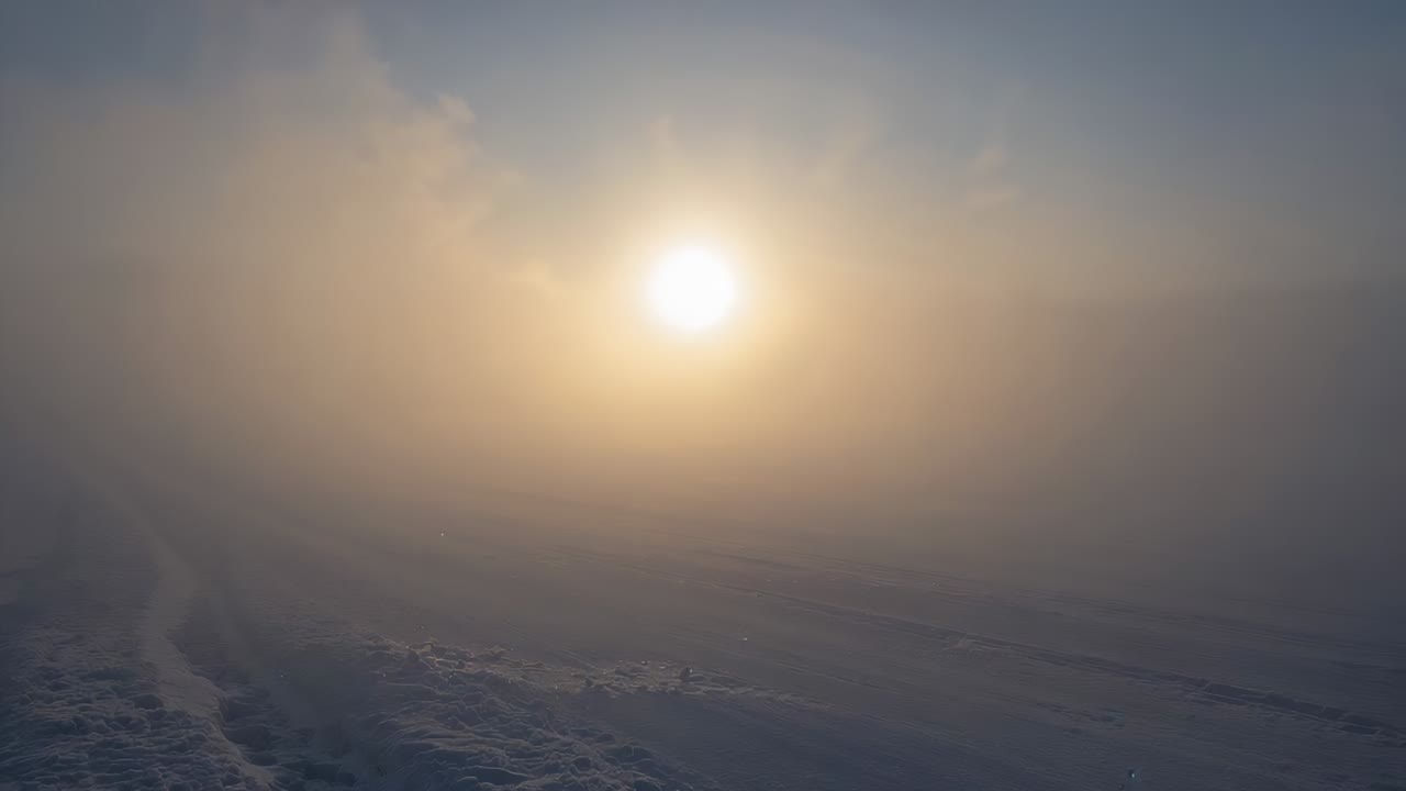 Shining low bright sun piercing band of haze over snowy plain, causing halo glow, tracks fading