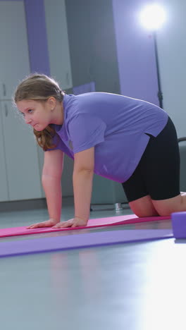 vista lateral de chicas jóvenes realizando ejercicios de flexibilidad de la columna vertebral en esteras en un gimnasio, con la chica de púrpura observando atentamente mientras la luz suave ilumina el fondo