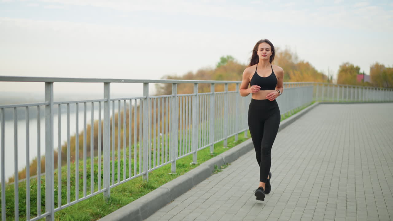 Young athlete in black sportswear running along path next to iron railing, embodying determination and energy, with a scenic view of the outdoors, showcasing athletic lifestyle and exercise