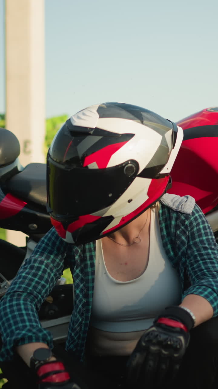una motociclista está sentada en la orilla de la carretera, las piernas cruzadas, tocando su casco en contemplación mientras está sentada al lado de una motocicleta roja