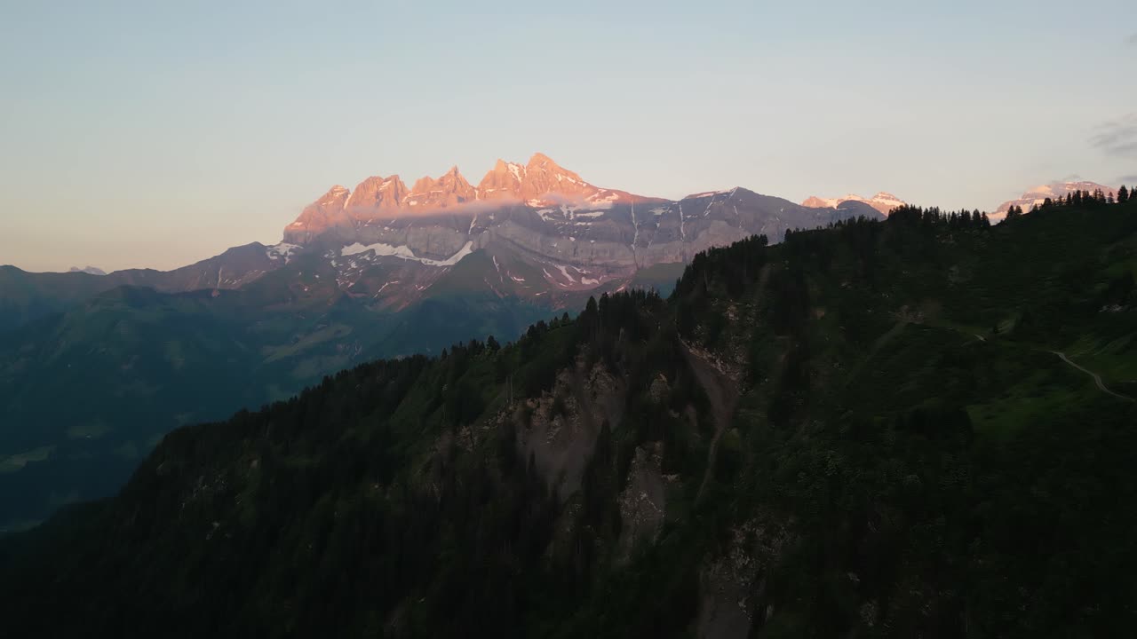 Beautiful Sunset drone flight in the Swiss Alps. Drone pans behind the tree line looking at the pink sunset on the peaks of the Dents du Midi in the Val d'Illiez of Canton Valais in Switzerland.