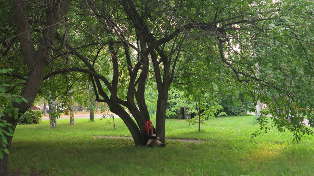solitary figure hiding behind low tree canopy in green park, knees drawn, shaded trunk sheltering subject, soft light filtering through leaves, contemplative stillness and introspective mood captured