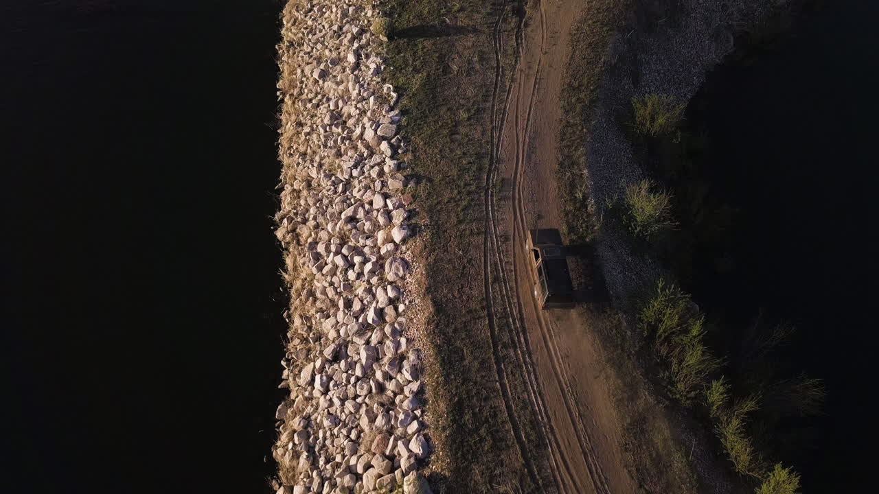 Vista da drone che segue un vecchio pick-up che percorre un frangiflutti dirigendosi verso la terraferma. Tramonto