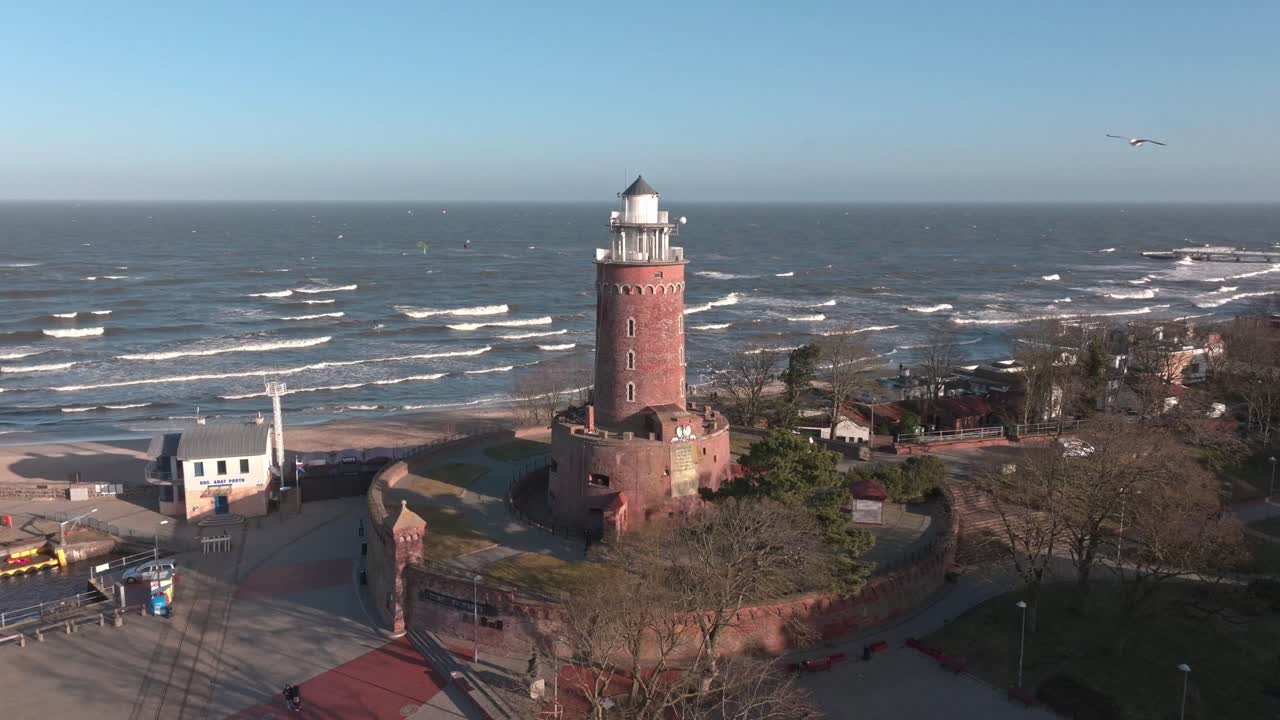 A lighthouse in the early spring morning against the backdrop of the Baltic Sea, with people surfing with the wind in the distance.